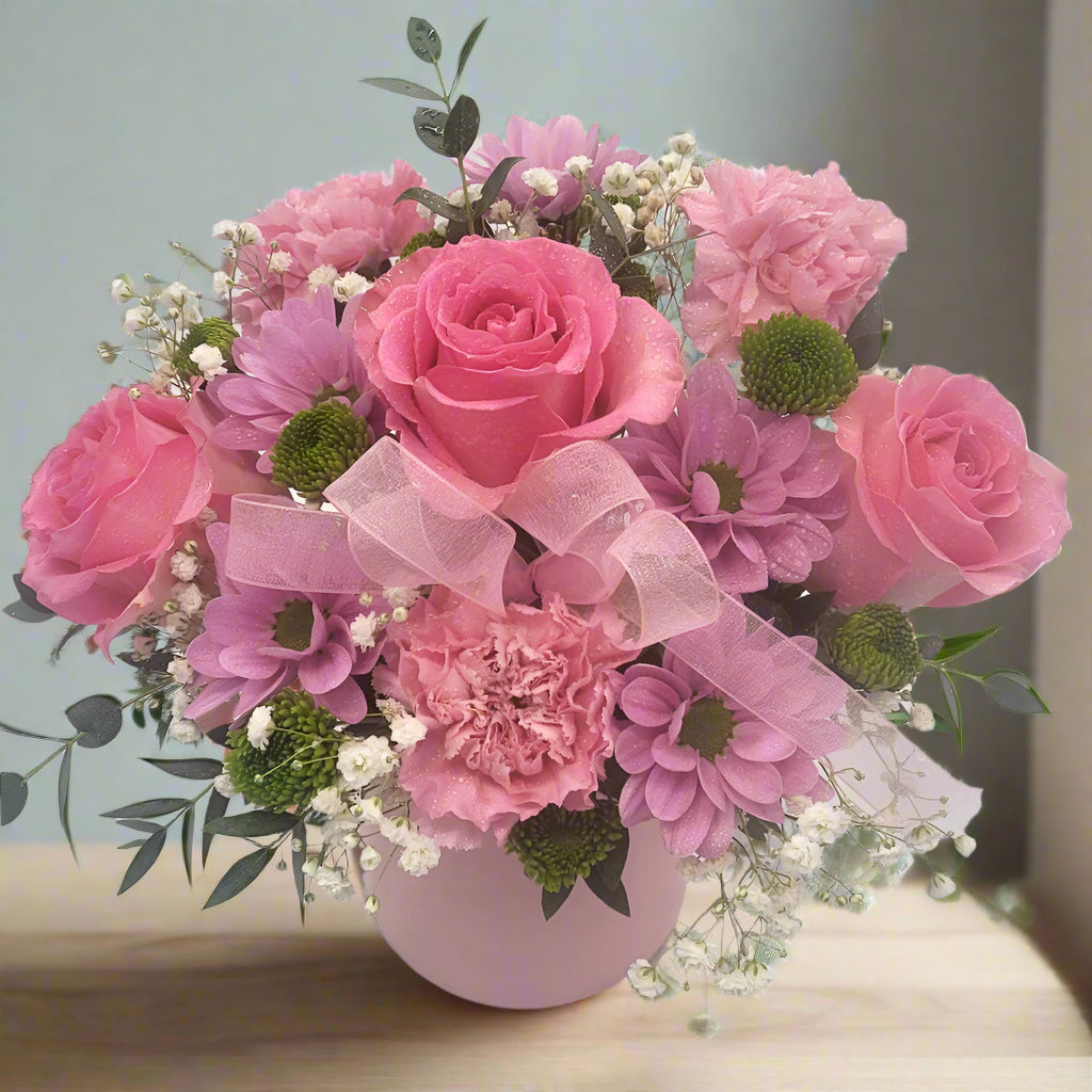 A bouquet of pink roses and carnations with greens and baby's breath, arranged in a round ceramic pot.