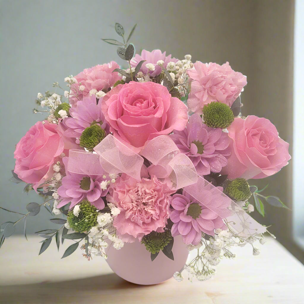 A bouquet of pink roses and carnations with greens and baby's breath, arranged in a round ceramic pot.