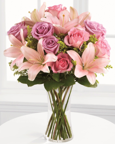 A sympathy bouquet with pink and lavender roses, lilies, and greenery in a glass vase.