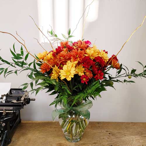 A large clear glass vase filled with a mix of fall-colored daisies, willow branches, and Italian ruscus.