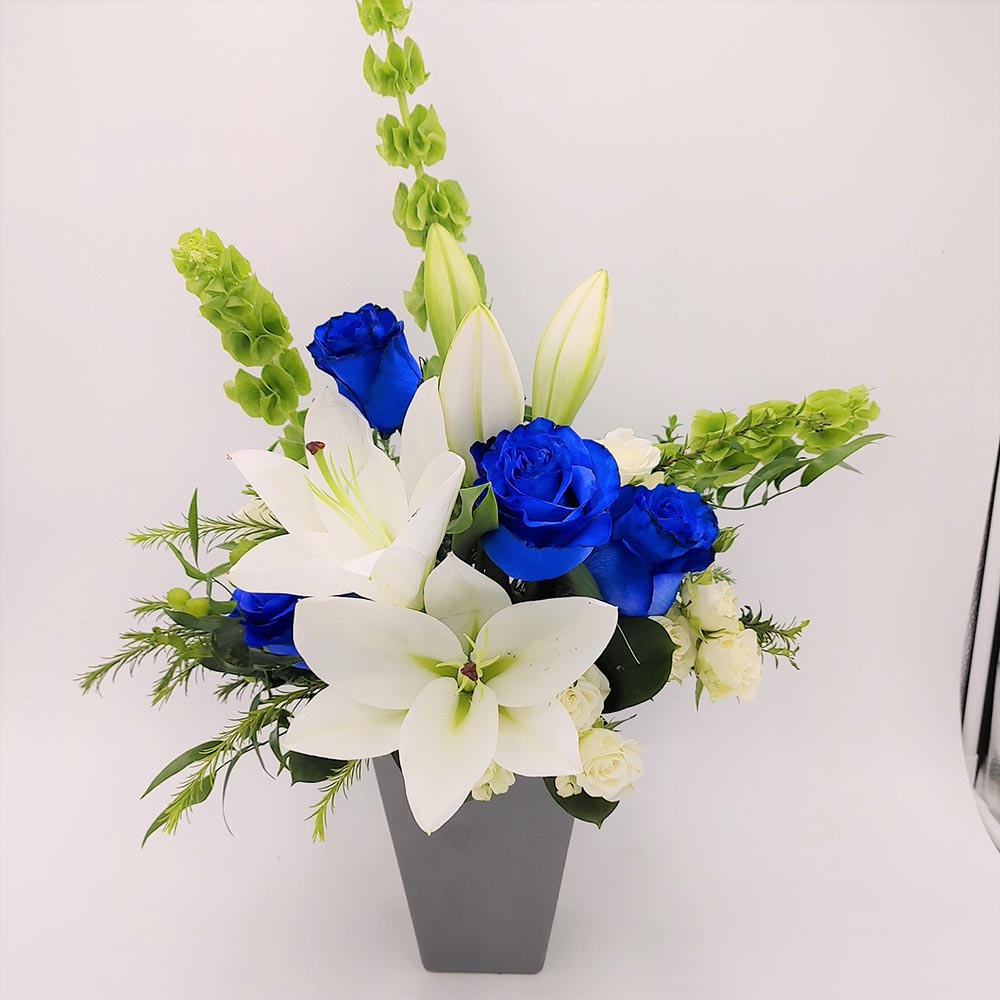Man of Steel Bouquet featuring blue roses, white lilies, spray roses & Bells of Ireland arranged with greens in a vase