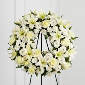 A white sympathy wreath with roses, lilies, mini carnations, and greenery, displayed on a wire easel for funerals and memorials