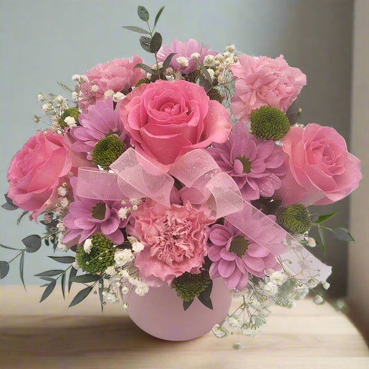 A bouquet of pink roses and carnations with greens and baby's breath, arranged in a round ceramic pot.
