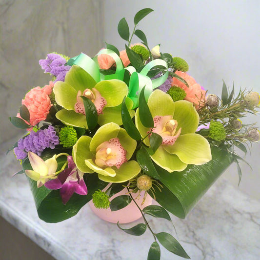 A floral arrangement featuring green Cymbidium Orchids in a ceramic vase, accented with seasonal flowers and greens.