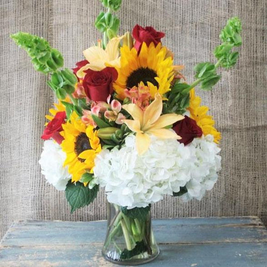 A bouquet of flowers including sunflowers, red roses, orange lilies, and spray roses with green foliage, arranged in a clear glass vase.
