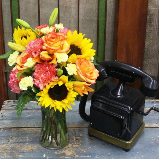 A clear glass vase containing a colorful bouquet of sunflowers, orange and yellow roses, and lush greenery, placed next to a vintage rotary dial phone on a wooden surface.