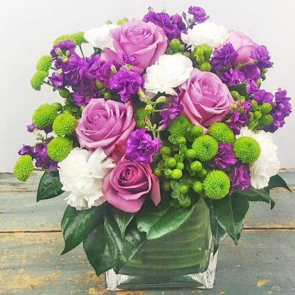 The Sweet Charm Bouquet with purple roses, carnations, purple stock & green button poms arranged in a square glass vase with lush greenery