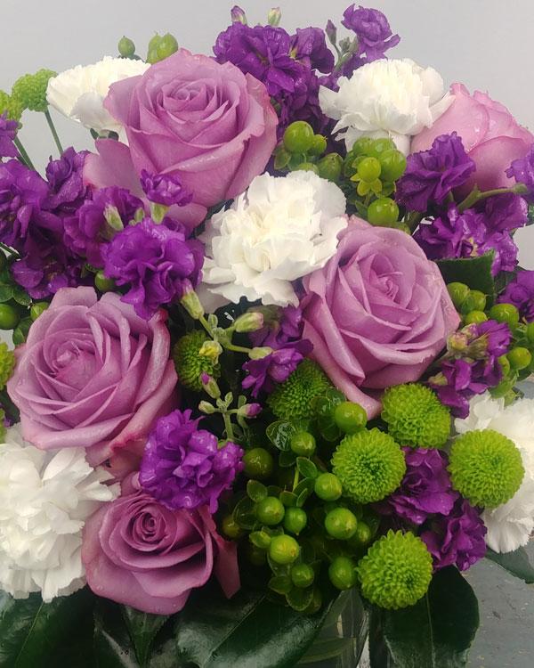 The Sweet Charm Bouquet with purple roses, carnations, purple stock & green button poms arranged in a square glass vase with lush greenery
