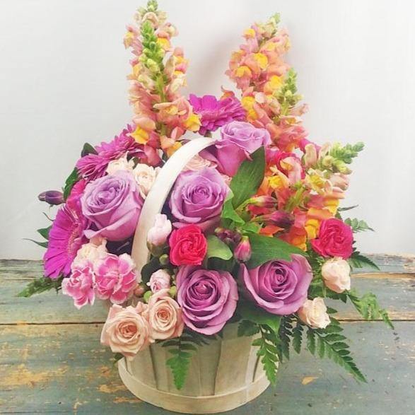 A floral basket arrangement with lavender roses, mini carnations, spray roses, and snapdragons, accented with lush greenery.