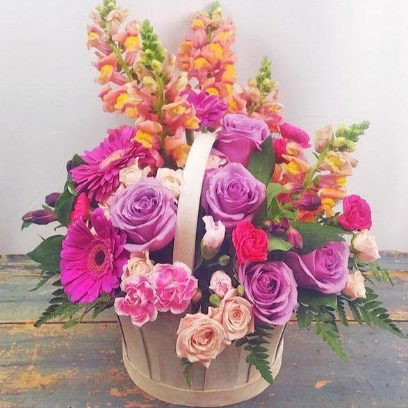 A floral basket arrangement with lavender roses, mini carnations, spray roses, and snapdragons, accented with lush greenery.