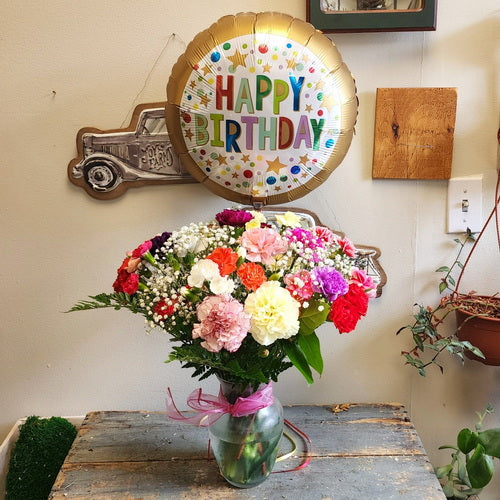 A bright birthday bouquet of carnations, baby’s breath, and greenery with a festive balloon in a clear glass vase.