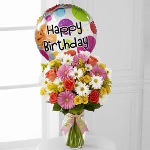 A vibrant birthday bouquet with coral roses, gerbera daisies, and a Happy Birthday Mylar balloon in a clear glass vase with ribbon.