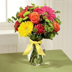 A bright bouquet of gerbera daisies, roses, carnations, and greenery in a clear glass vase with a yellow satin ribbon.