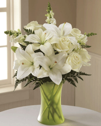 A bouquet of white roses, lilies, and snapdragons in a green glass vase for sympathy.