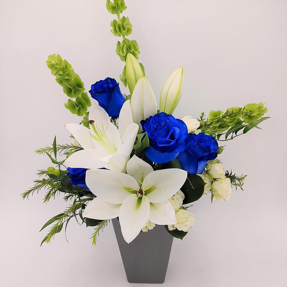 Man of Steel Bouquet featuring blue roses, white lilies, spray roses & Bells of Ireland arranged with greens in a vase