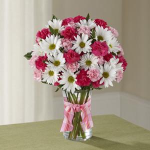 A bouquet of pink carnations, white daisies, and greenery in a clear glass vase with a pink ribbon.